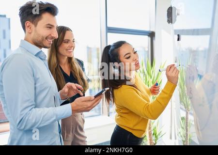 Businesswoman writing on whiteboard with colleagues in background at office Stock Photo