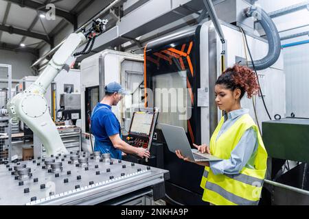 Young engineer using laptop with colleague operating machine in factory Stock Photo