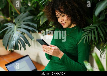 Smiling freelancer text messaging through smart phone surrounded by plants Stock Photo