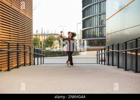 Young woman taking selfie and walking on footpath amidst buildings Stock Photo