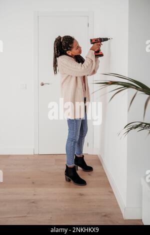 Mature woman using drill machine on wall at home Stock Photo
