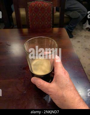 Closeup of nearly empty glass of beer with layer of foam and human hand on a wooden table in a restaurant. Stock Photo