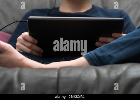Digital tablet computer in male hands, from below view closeup. Young guy sitting on sofa play videogame on handheld gadget. Modern technology, tablet Stock Photo