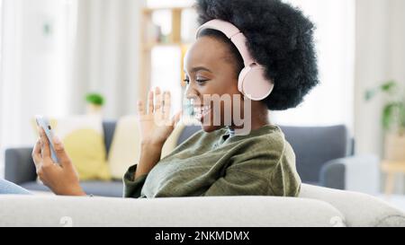 Cheerful african afro woman using phone and headphones and waving while on a video call with friends. Remote student using mobile app and talking to Stock Photo