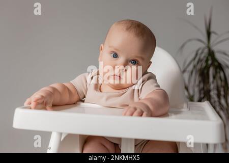 chubby baby in a white bodysuit sits in a white high chair for feeding. High quality photo Stock Photo
