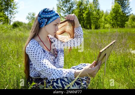 Woman sitting on grass looking at her mirror image. Portrait of attractive girl in dress on a sunny summer day Stock Photo