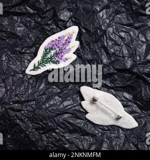 The brooch is embroidered with threads on white felt in the form of a heather, next to it lies its back side with a pin.  Against the background of cr Stock Photo
