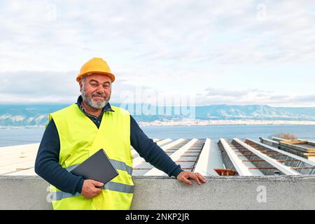 Portrait of smiling middle aged bearded supervisor in hardhat and safety vest with tablet on building site. Structural engineer or architect monitors Stock Photo