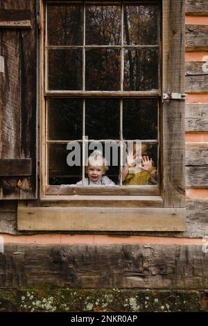 Two children looking through window of wooden hut towards camera Stock Photo