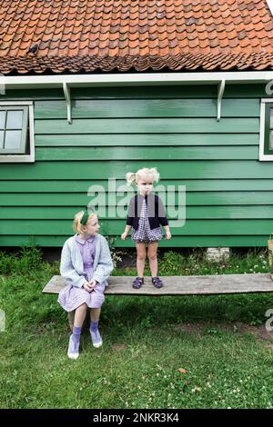Two girls outside wooden house, one standing and one sitting on bench Stock Photo
