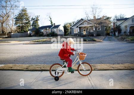 Girl wearing red coat riding bicycle along sidewalk Stock Photo