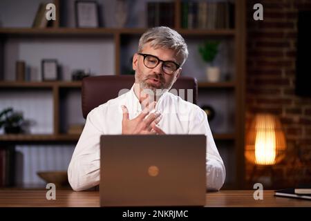 Gray-haired manager in glasses uses a laptop, conducts a video conference in the office. Middle aged businessman participating in online webinar Stock Photo