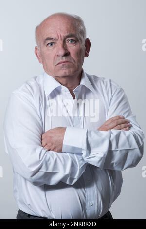 Confident senior man in white shirt crossing hands on chest and looking at camera while standing against gray background. Self confident senior Stock Photo