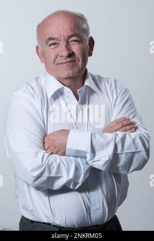 Confident senior man in white shirt crossing hands on chest and looking at camera while standing against gray background. Self confident senior Stock Photo