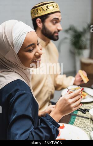 Smiling african american woman in hijab holding tea glass near blurred husband during ramadan at home,stock image Stock Photo