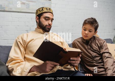 Muslim man reading book near son on couch at home,stock image Stock Photo