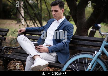 Young elegant businessman sitting on a bench in the park, using smartphone. Stock Photo