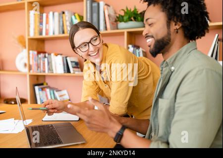Pair of friendly colleagues work in office. Young businessman explains to his female colleague something using a laptop, they are looking on the screen Stock Photo