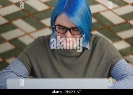 Portrait of white woman with dyed blue hair working on computer at home. Close up photo of female person in glasses studying online with a laptop Stock Photo
