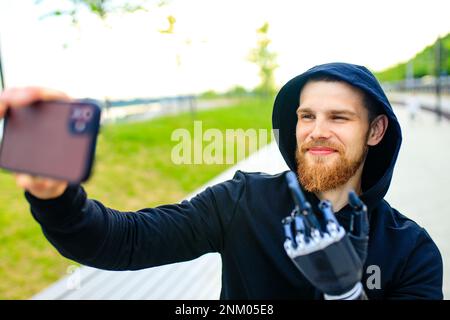 young bearded man with metal arm prosthetic outdoor taking selfie photos on smartphone camera Stock Photo