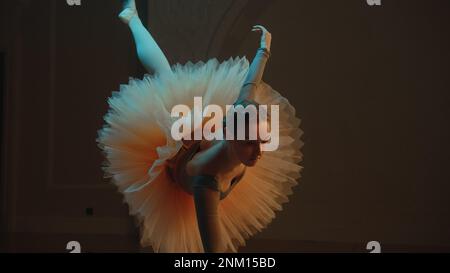 Cinematic shot of graceful ballerina in ballet tutu performing in dim opera lobby and practicing gymnastic movement. Female ballet dancer on choreography rehearsal in theater. Classical ballet art. Stock Photo