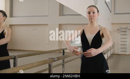 Graceful adult ballerina in training bodysuit rehearses near ballet barre and practices classical ballet movements in dance studio. Female ballet dancer on choreography rehearsal. Ballet dance school. Stock Photo