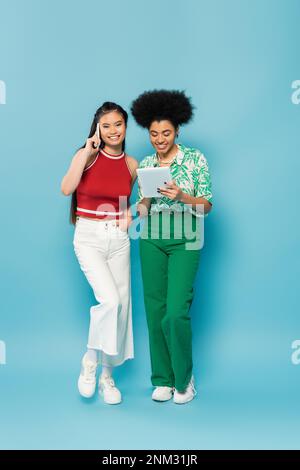 full length of happy asian woman talking on smartphone near african american friend using digital tablet on blue background,stock image Stock Photo