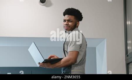 Two multi ethnic installers in uniform sets up security cameras in office corridor using laptop. Upset African American worker looks at camera. Concept of surveillance systems and privacy. Stock Photo