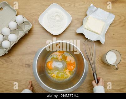 Kid's hands with mixing whisk. Eggs, sugar, butter, flour. Cooking ingredients in bowl, ready for mixing dough. Cookbook. Stock Photo
