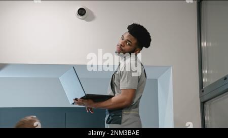 Two multi ethnic installers in uniform sets up security cameras in office corridor using laptop. Upset African American worker looks at camera. Concept of surveillance systems and privacy. Stock Photo
