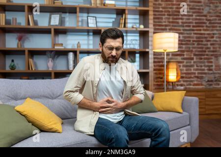 A young man feels severe stomach pain, spasms, poisoning, discomfort. Sitting on the sofa at home and holding his stomach. Stock Photo