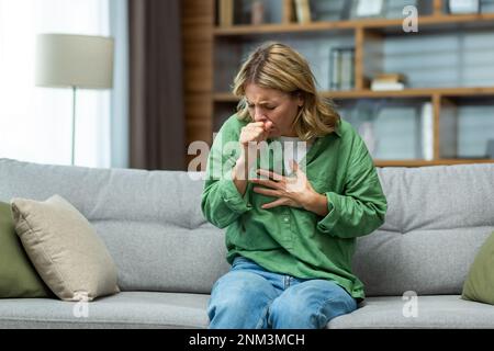 Senior beautiful woman sitting sick on sofa at home. He coughs, covers his mouth with his hand, holds his chest. Feels pain, suffers from asthma, allergies, flu, cold. Stock Photo