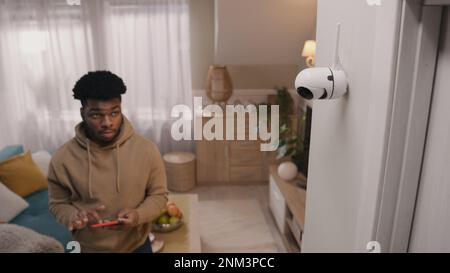 African American man completes installation of security camera. Man sets up angle of CCTV camera at home and rotates it using smartphone and app. Concept of monitoring system, safety and privacy. Stock Photo