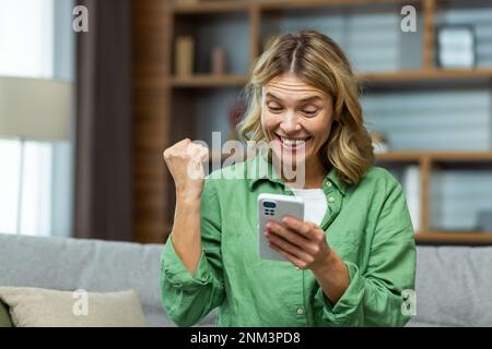 Close-up photo. A happy senior woman is sitting on the sofa at home, holding the phone in her hands. Shows a victorious yes gesture with his hand. Received good news, financial reward, prize, lottery. Stock Photo