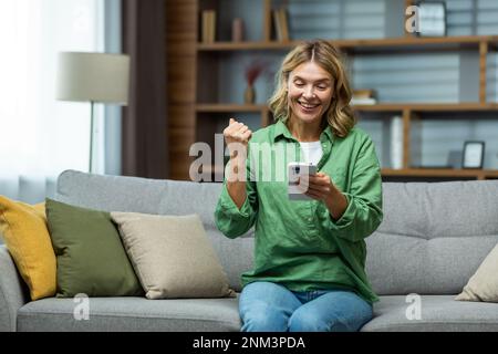 A happy senior woman is sitting on the sofa at home, holding the phone in her hands. Shows a victorious yes gesture with his hand. Stock Photo