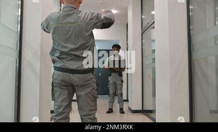 Two diverse men in uniform walk corridor and discuss CCTV cameras installation in business office. Installers set up security cameras using computer program on laptop. Concept of surveillance system. Stock Photo