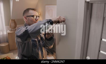 Man in uniform puts cable in drilled hole. Electrician removes insulation from wire with professional tool in apartment. Repair work on electricity. Installation of the Internet or security cameras. Stock Photo