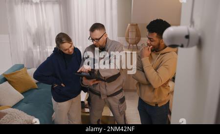 Installer sets up security camera in apartment using digital tablet. Man shows to multi cultural couple how to check motion sensor of CCTV camera in app. Tracking system, safety and privacy concept. Stock Photo