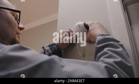 Installer in uniform puts security camera on wall fastening and connects it to system with cable. Man installs cameras in house. Concept of CCTV cameras, monitoring, safety and privacy. Stock Photo