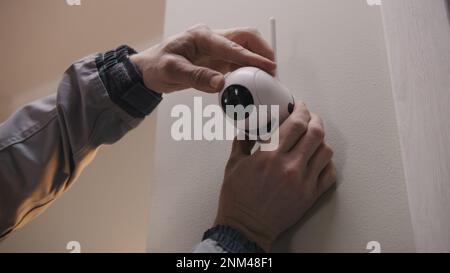 Installer in uniform puts security camera on wall fastening and connects it to system with cable. Man installs cameras in house. Concept of CCTV cameras, monitoring, safety and privacy. Stock Photo