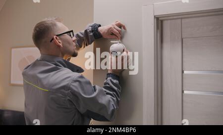 Installer in uniform puts security camera on wall fastening and connects it to system with cable. Man installs cameras in house. Concept of CCTV cameras, monitoring, safety and privacy. Stock Photo