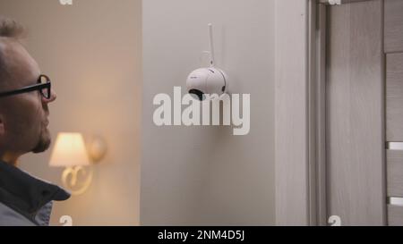 Installer in uniform puts security camera on wall fastening and connects it to system with cable. Man installs cameras in house. Concept of CCTV cameras, monitoring, safety and privacy. Stock Photo