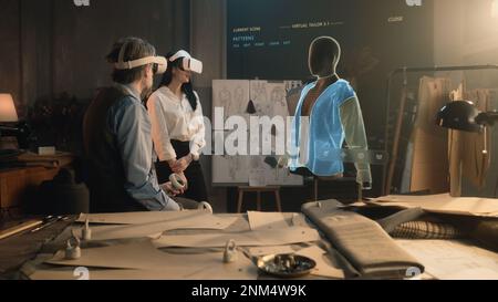 Tailor and female customer put VR goggles in atelier workshop. 3D hologram of jacket pattern. Virtual interface menu of program for designing and modeling clothes. Technologies of augmented reality. Stock Photo