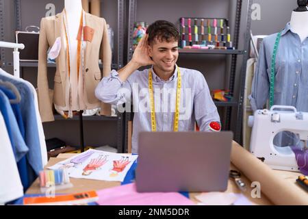 Young hispanic man dressmaker designer using laptop smiling with hand over ear listening and hearing to rumor or gossip. deafness concept. Stock Photo