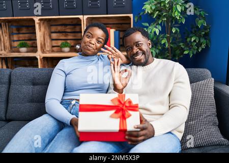 Young african american couple holding present doing ok sign with fingers, smiling friendly gesturing excellent symbol Stock Photo