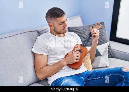 Young hispanic man playing ukelele sitting on sofa at home Stock Photo