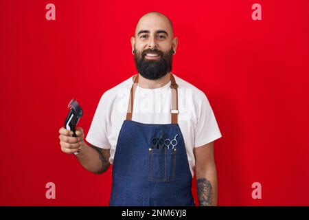 Young hispanic man with beard and tattoos wearing barber apron holding razor with a happy and cool smile on face. lucky person. Stock Photo