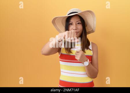 Middle age chinese woman wearing summer hat over yellow background punching fist to fight, aggressive and angry attack, threat and violence Stock Photo