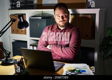 Plus size hispanic man with beard working at the office at night smiling looking to the side and staring away thinking. Stock Photo