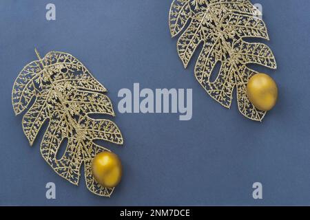 Gold eggs on monstera leaves from wire on blue background. Stock Photo